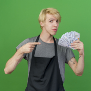 professional barber man in apron holding cash pointing with index finger at money looking confident standing over green background